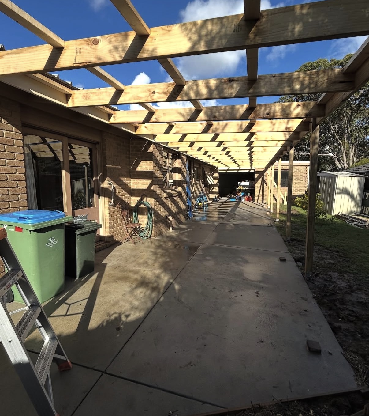Large timber pergola frame under construction over concrete driveway beside brick house